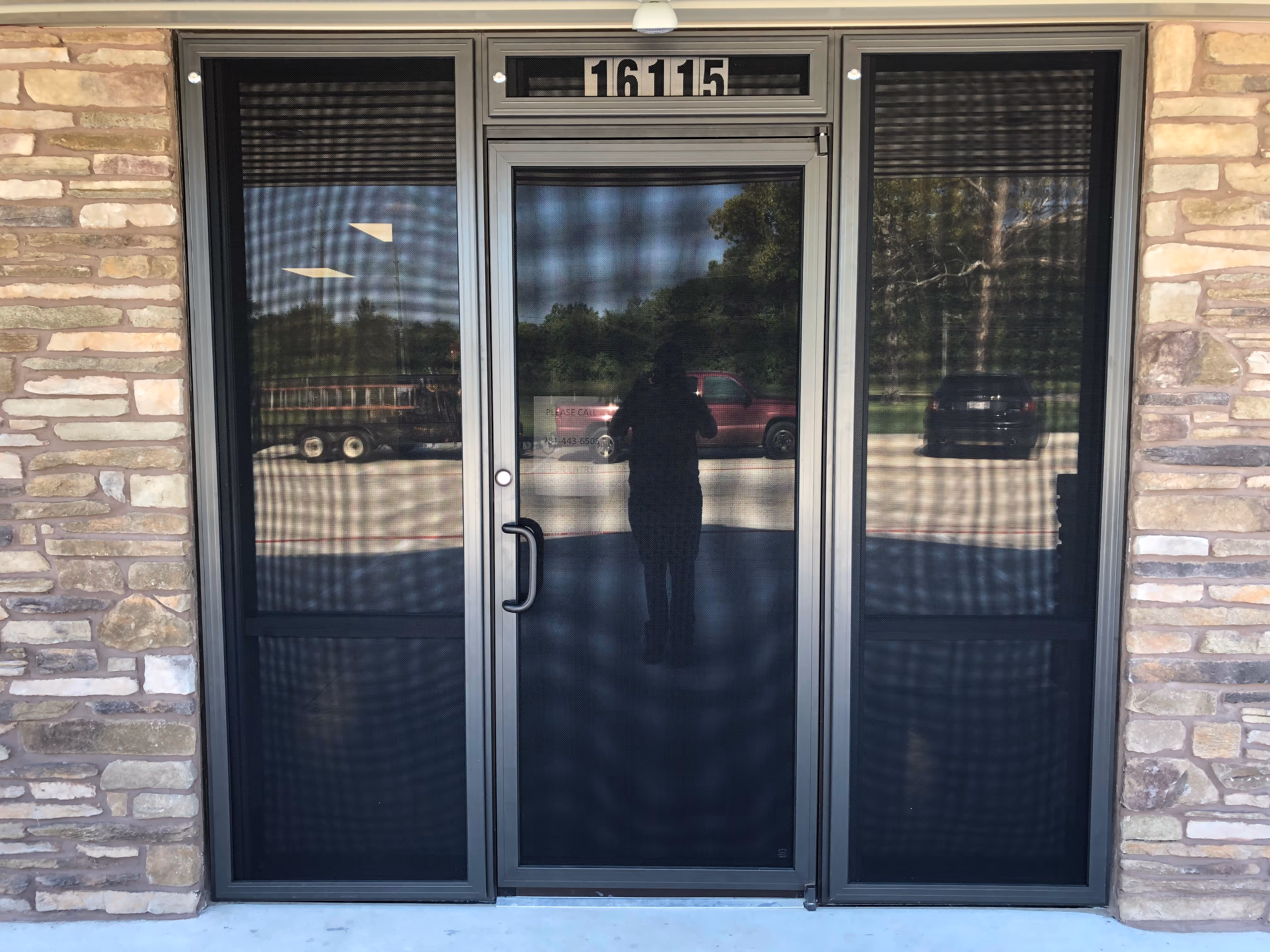 Front entrance with three gray-framed mesh screen doors set in a stone wall, showing a reflection of a person taking a photo and cars parked outside.