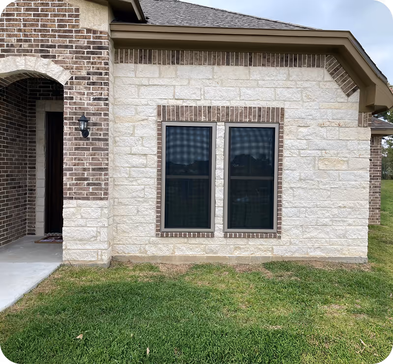 Exterior house wall with light-colored stone bricks, two narrow windows with dark frames, and part of a covered entrance with dark bricks and a black wall-mounted lamp.