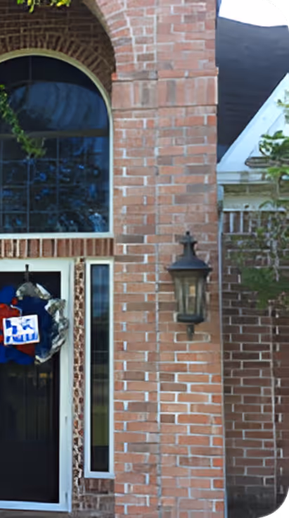 Red brick house entrance with a black door, side window, arched transom window above, a decorative wreath on the door, and a wall-mounted lantern light on the brick wall.
