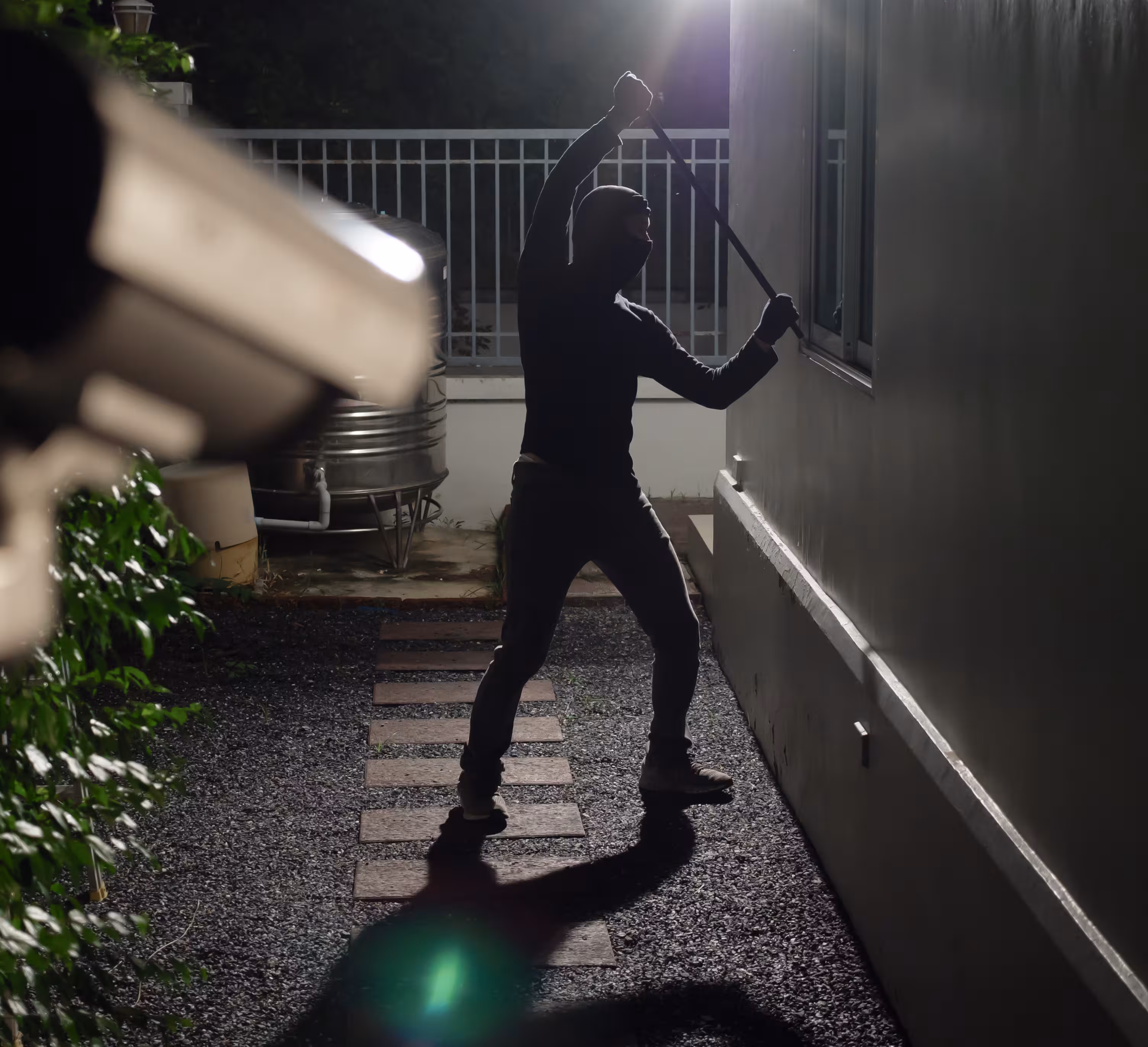Silhouette of a masked person breaking a window with a crowbar at night near a building.