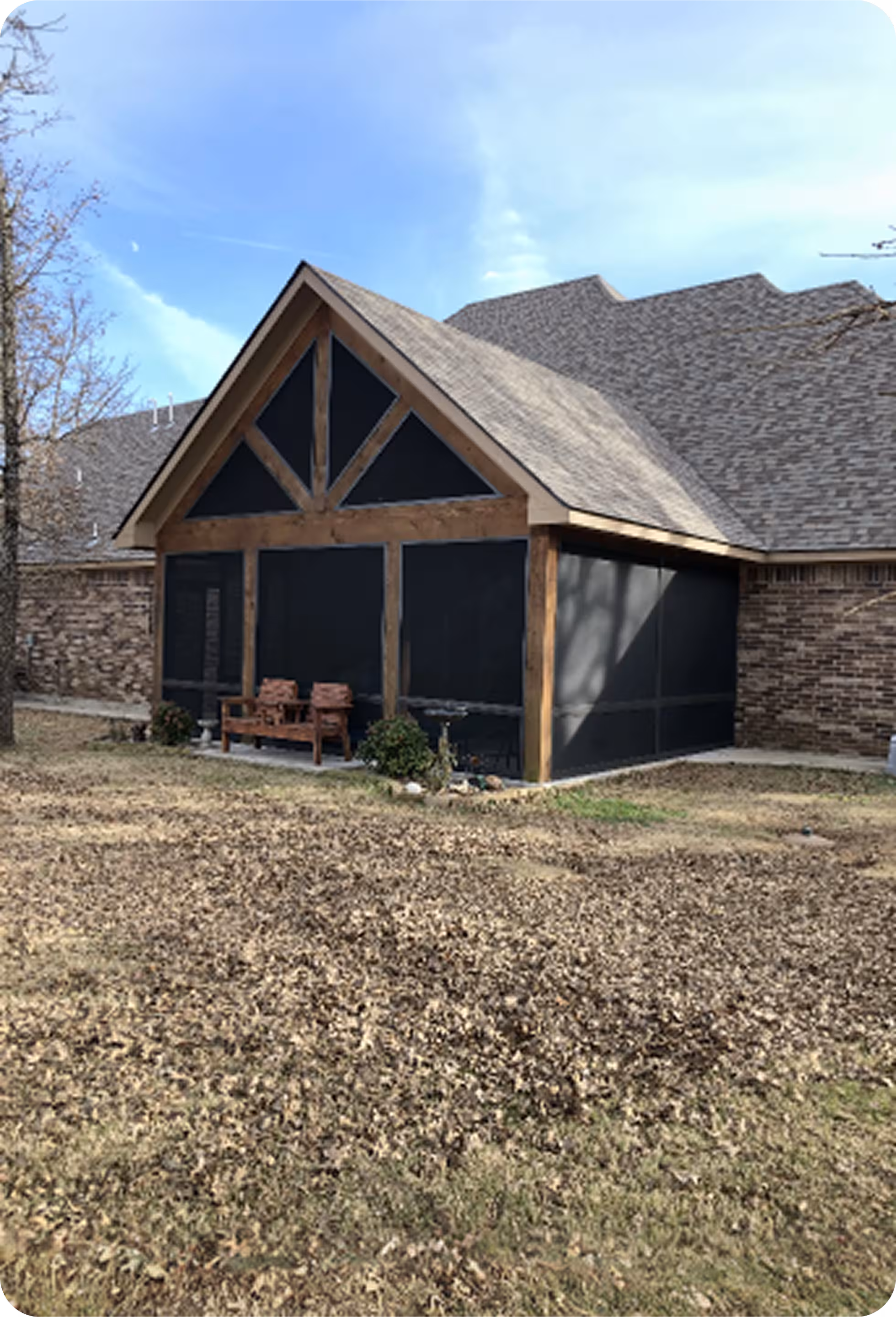 Back view of a brick house with a gable-roofed screened porch and wooden bench on a yard covered with dry leaves.