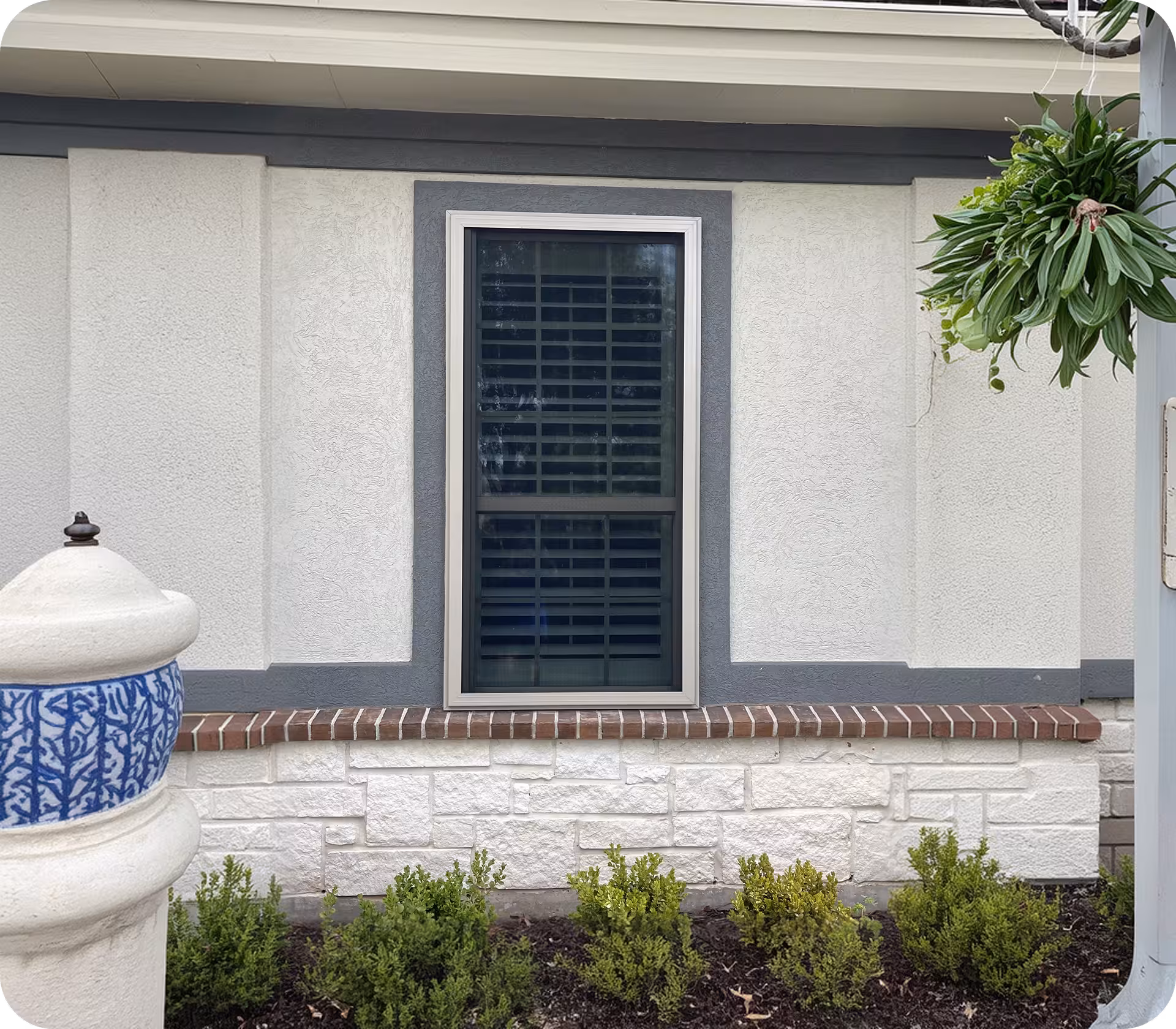 Exterior wall with a closed window featuring horizontal blinds, small shrubs planted at the base, and a decorative white and blue post on the left.