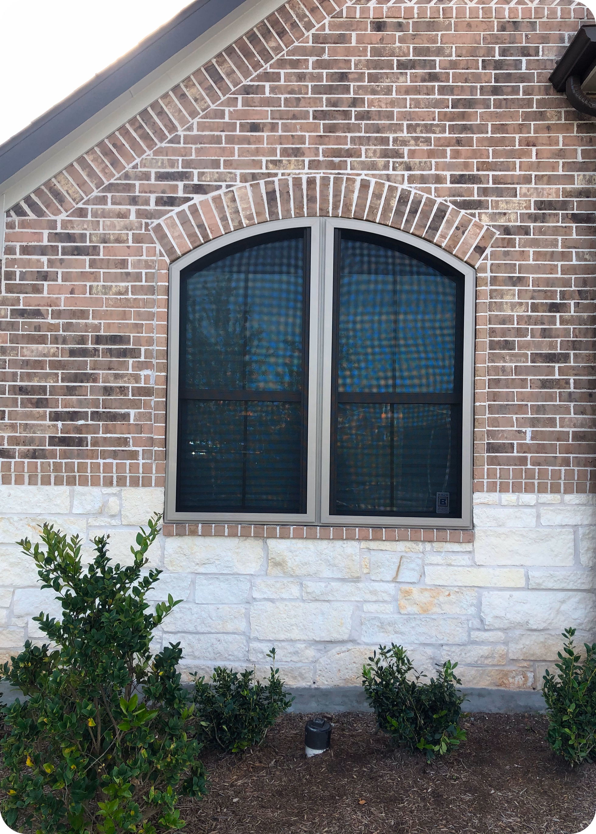 Arched window with brown frame on a brick and white stone house wall, with green bushes and mulch below.