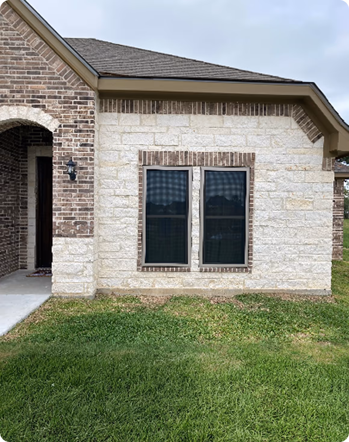 Exterior of a house with light-colored stone walls, two dark windows with brick trim, and a small covered entrance on the left.