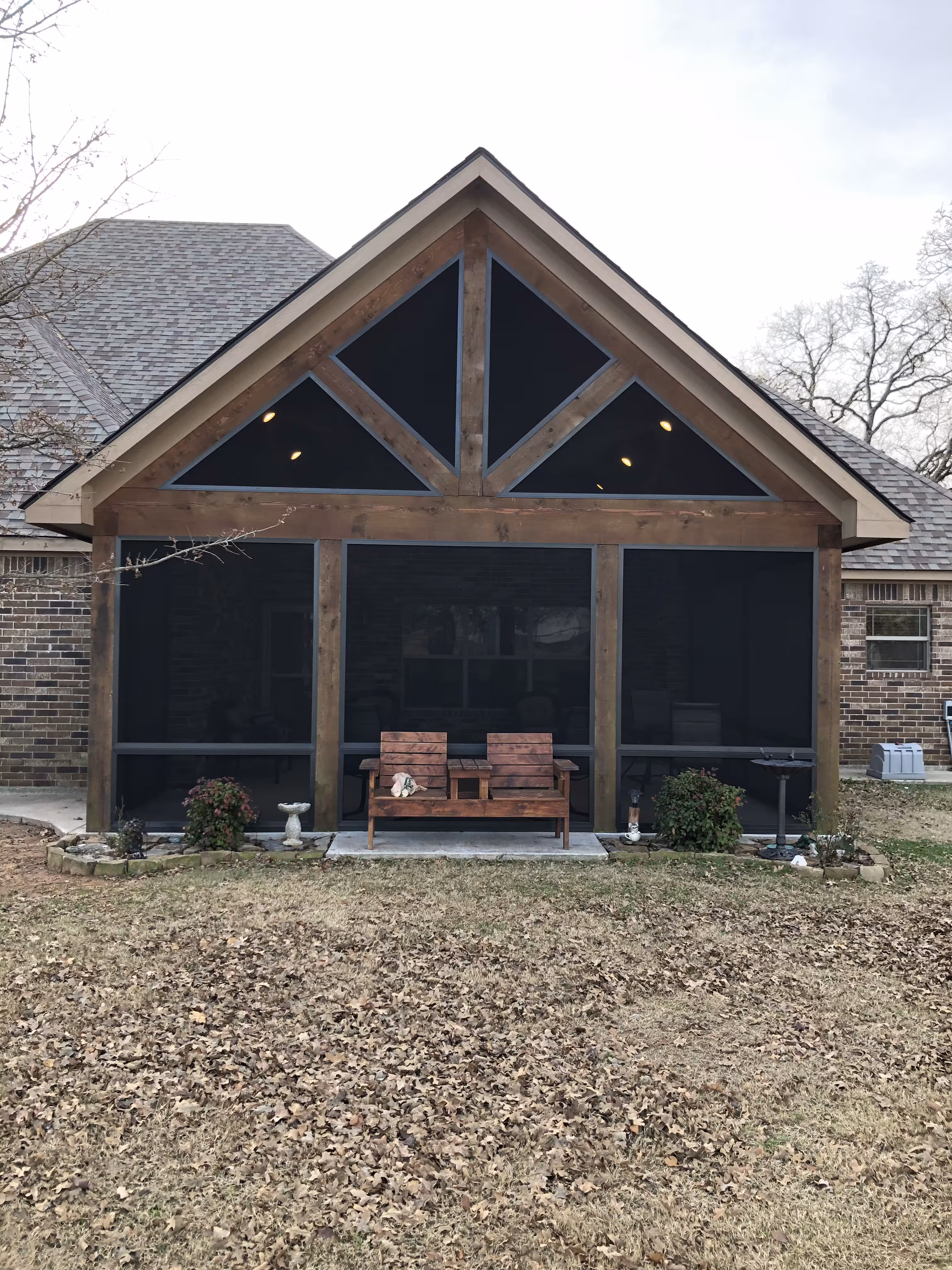 Backyard view of a screened-in porch with wooden framing, two chairs with a small table in between, and dry leaves covering the lawn.