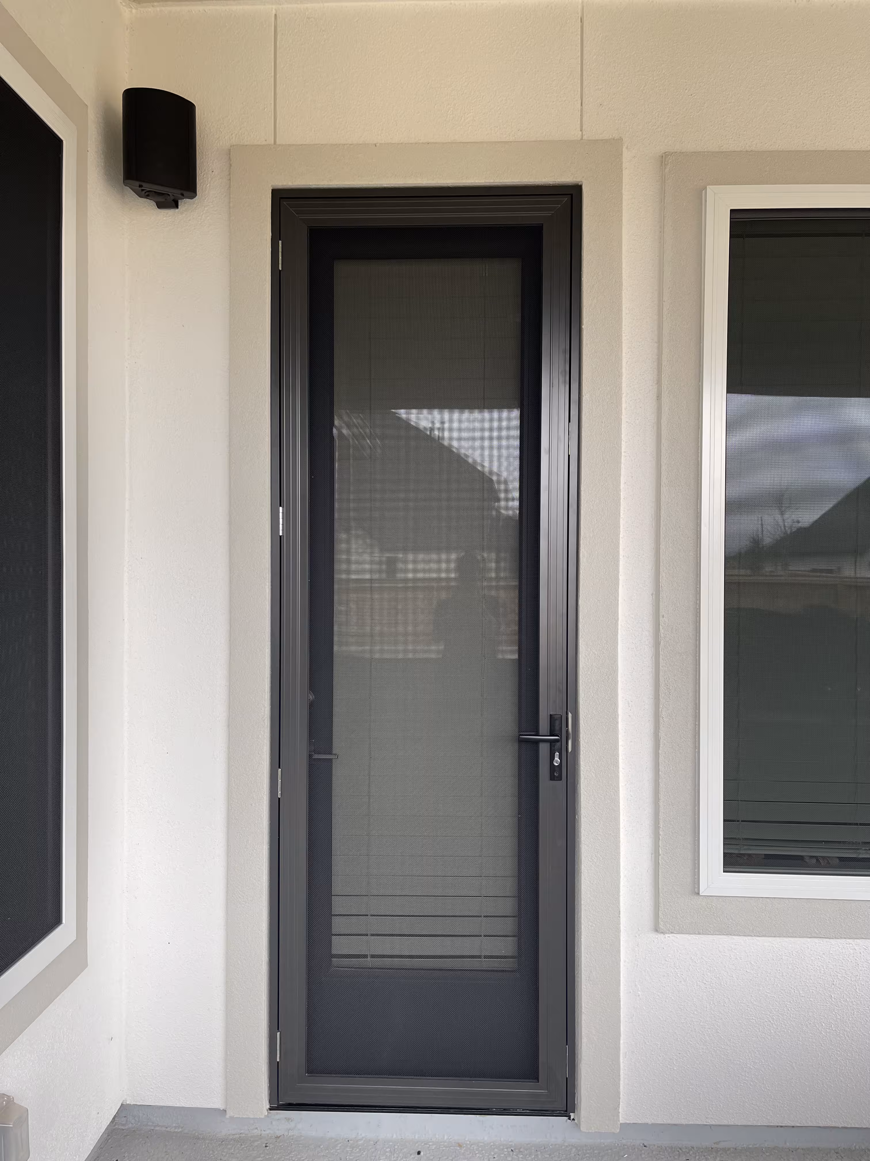 Black framed screen door with handle on a beige stucco wall, flanked by two windows with blinds and a black outdoor light fixture above the left window.
