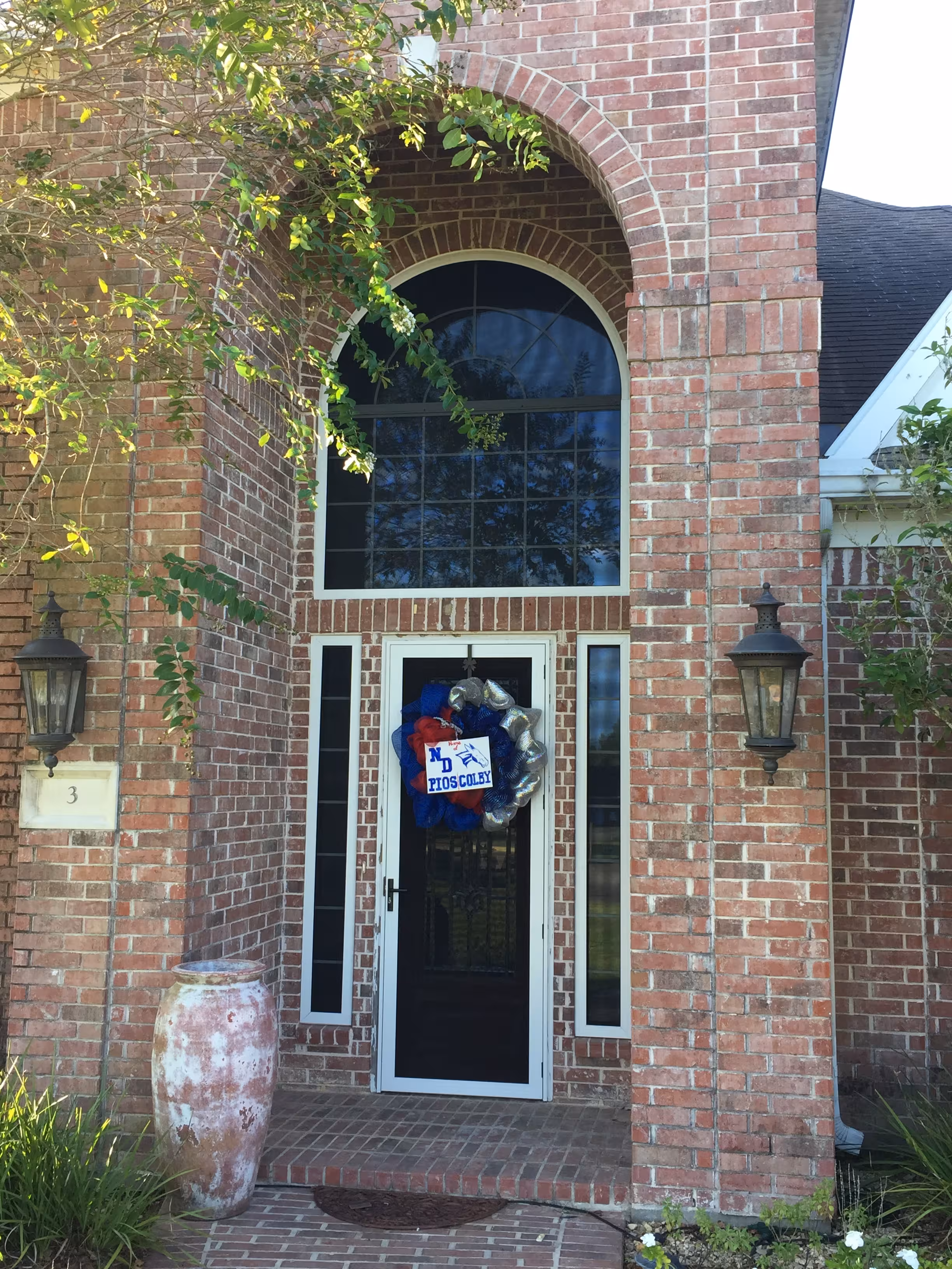 Brick house entrance with a tall arched window, a glass door decorated with a colorful wreath, two black lantern-style wall lights, and a large weathered ceramic vase.