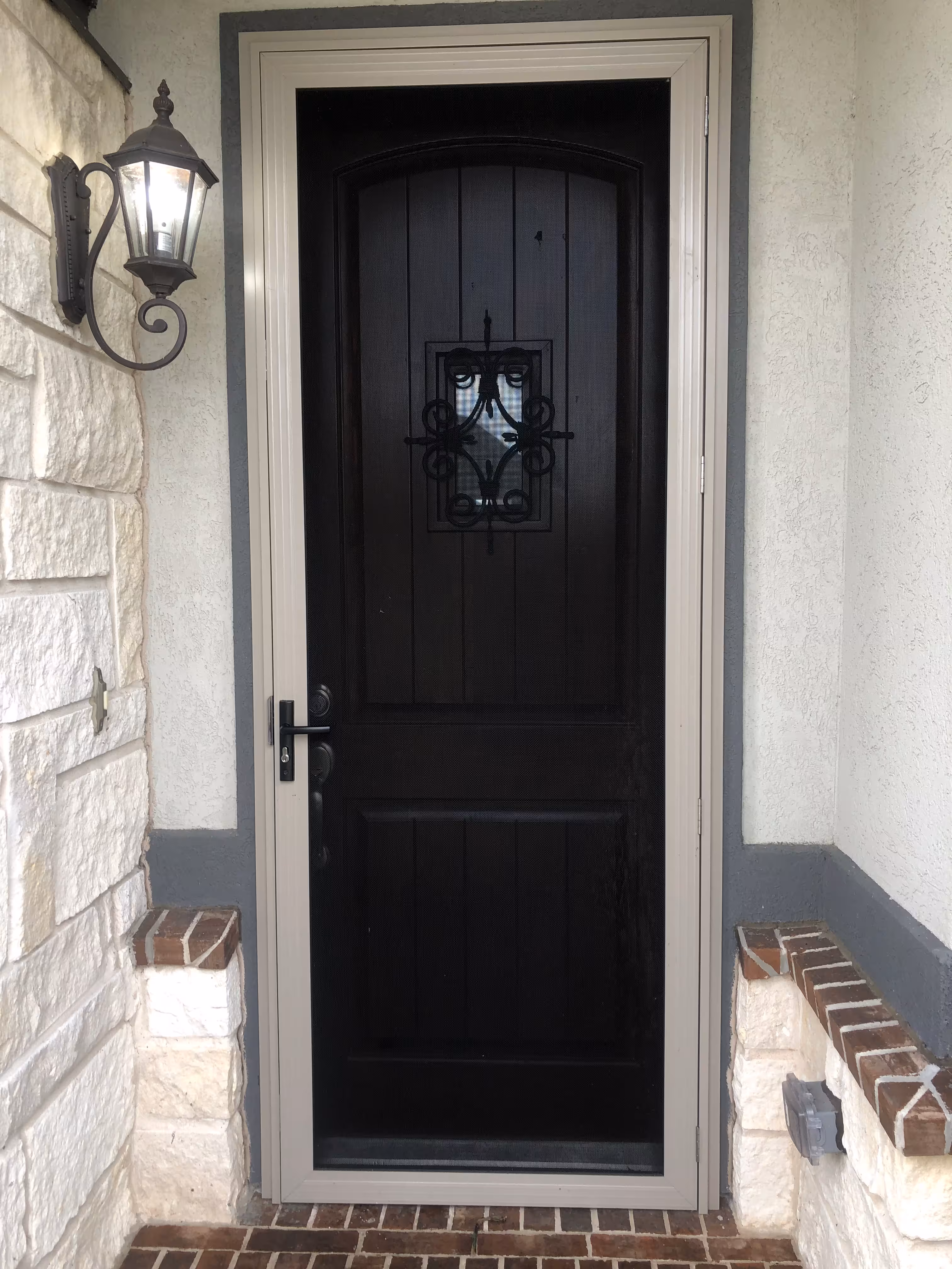 Black wooden front door with a small decorative window and metal grille, framed by beige trim and surrounded by light stone and stucco walls with a lit wall lantern on the left.