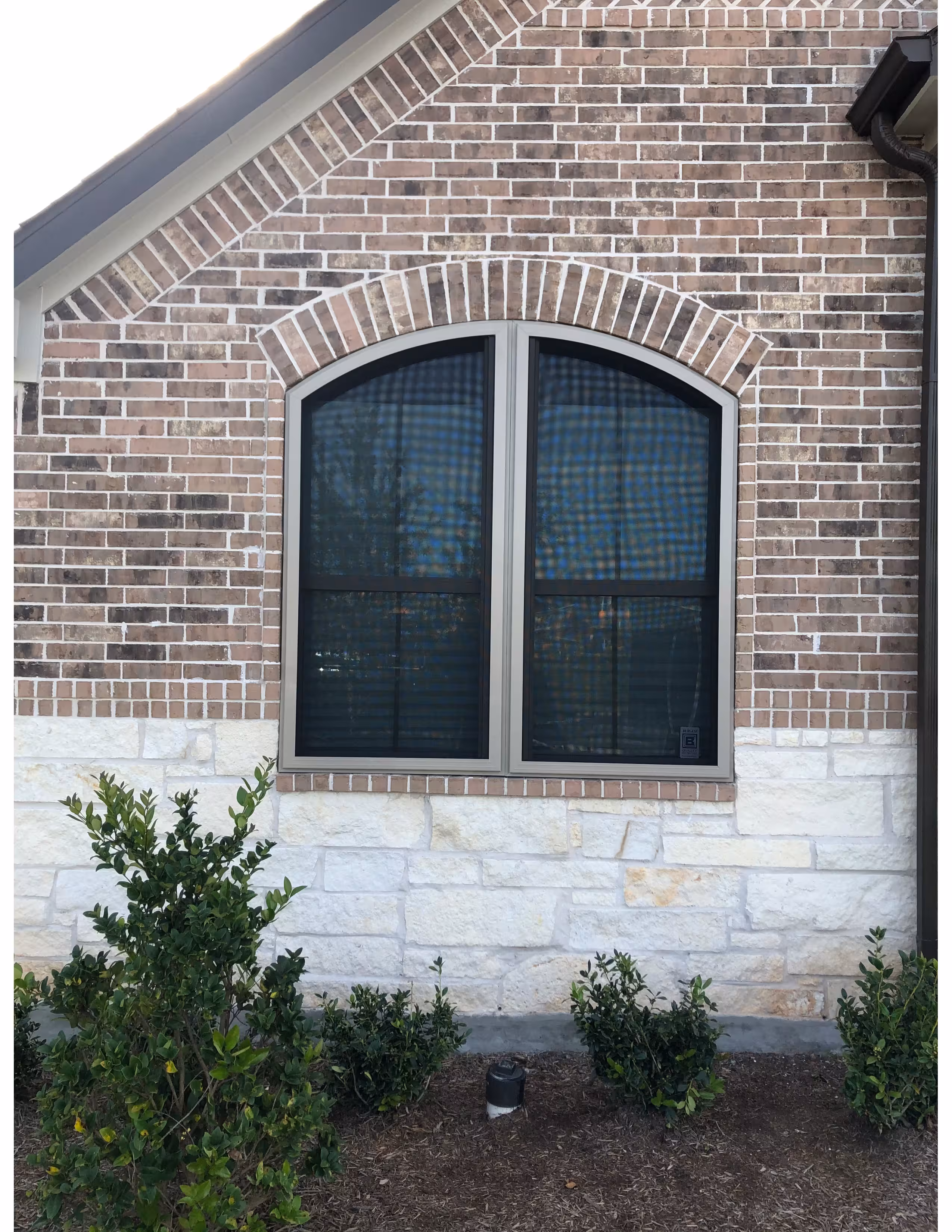 Arched double window with beige frame set in a brick and stone exterior wall, surrounded by green shrubs.