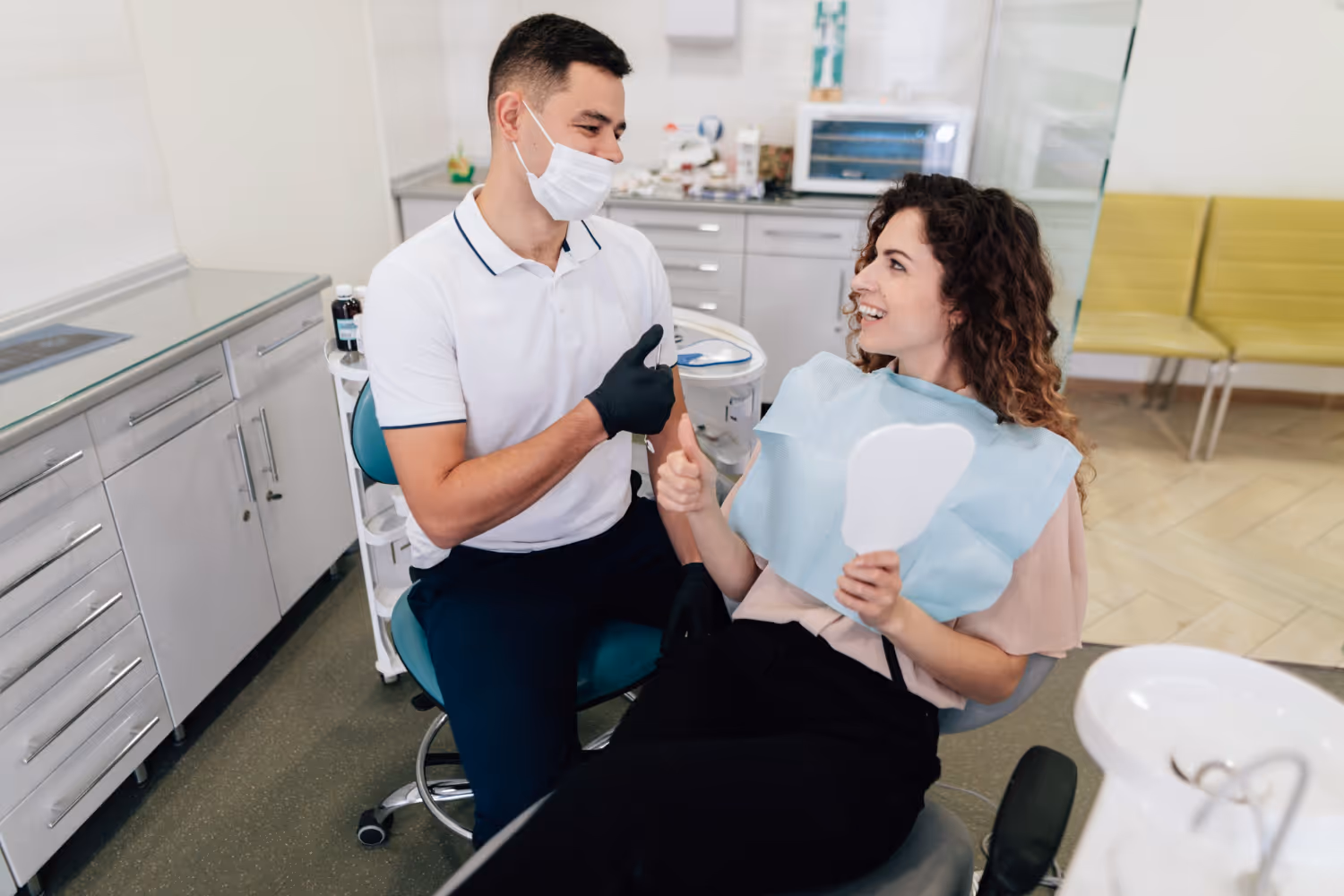 Female dental patient smiling and giving thumbs up while holding a hand mirror, with male dentist wearing mask and gloves giving thumbs up in a dental clinic.