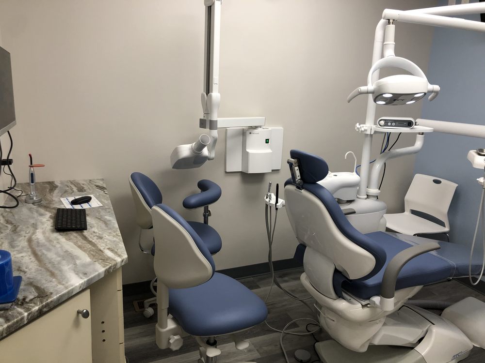 Modern dental exam room with dental chair, stools, overhead light, and countertop with keyboard and dental tools.