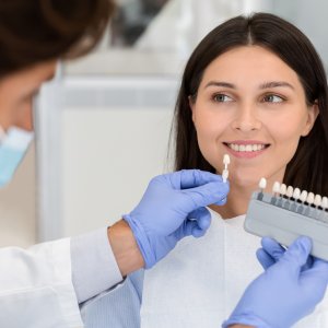 Dentist with blue gloves comparing a dental shade guide to a smiling woman's teeth for color matching.