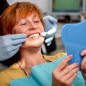 Smiling woman in dental chair looking at her teeth in a handheld mirror while a dentist wearing blue gloves examines her mouth.