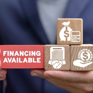Hand holding wooden blocks with financing icons and a red block reading 'Financing Available'.