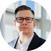 Man with short dark hair and glasses wearing a white shirt and dark blazer in an indoor setting.