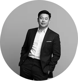 Confident man in a dark suit with hands in pockets standing against a plain background.