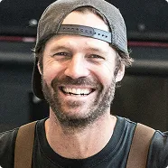 Smiling man with beard wearing a backward baseball cap and black shirt.