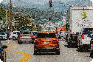 Traffic on a city street with cars and trucks stopped at a red light against a backdrop of hills.
