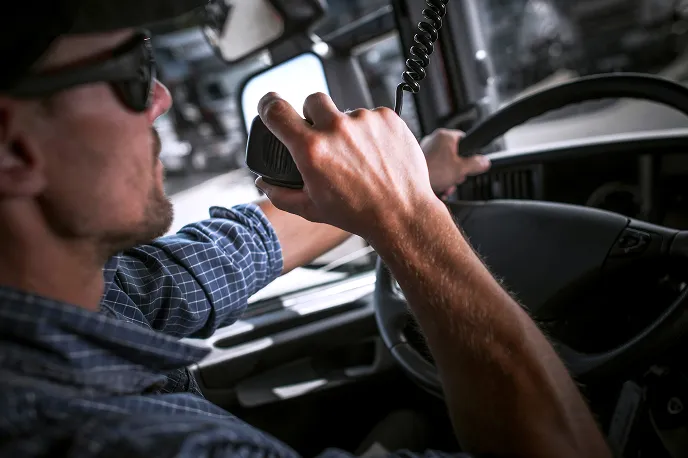 Man wearing sunglasses and a checkered shirt speaking into a handheld radio while driving a vehicle.