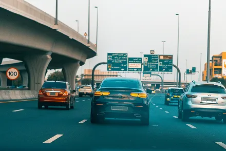 View of a highway with multiple cars driving, overhead road signs in Arabic and English, and a speed limit sign of 100 km/h.