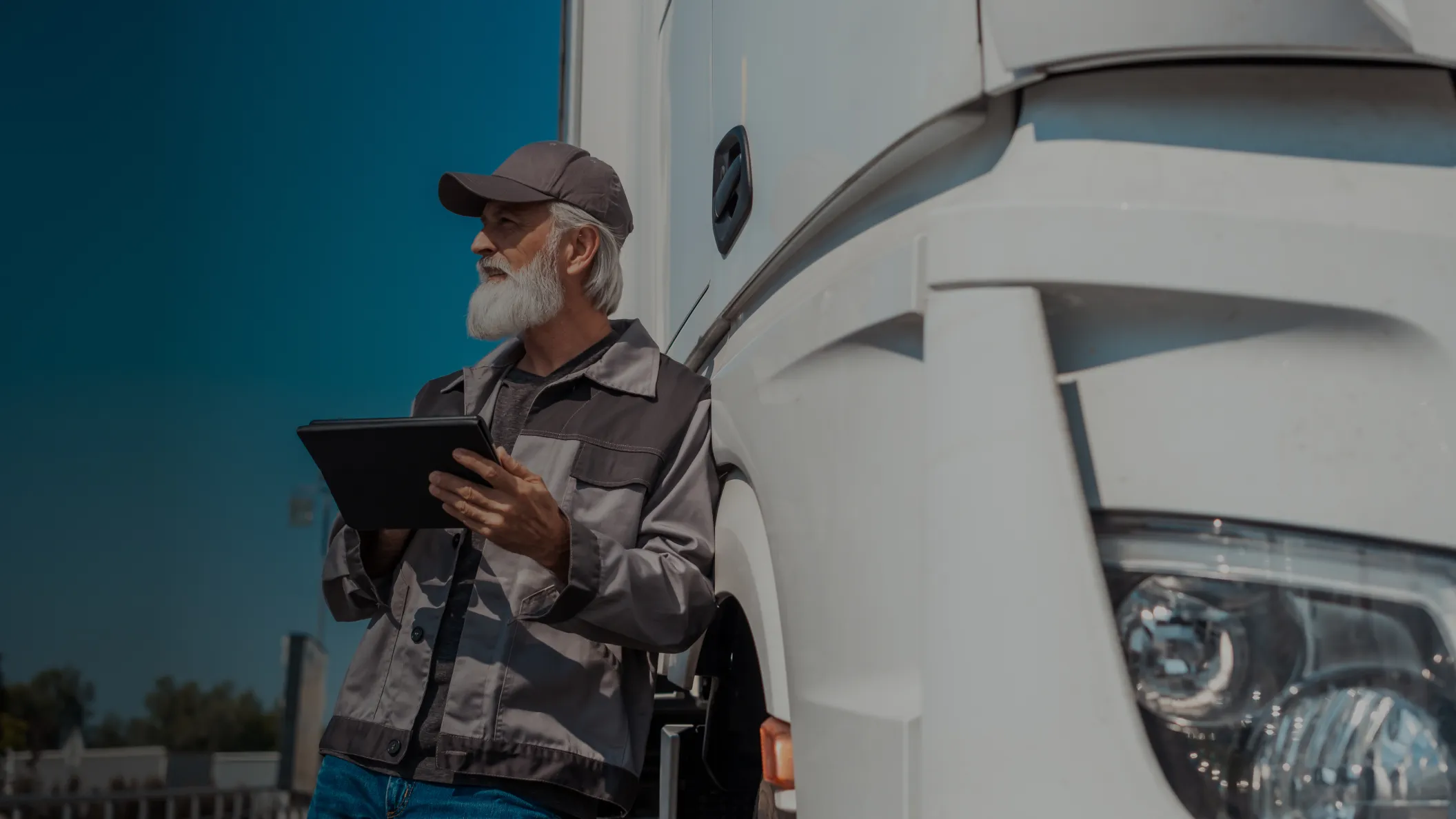 Bearded older man in work jacket and cap standing beside a white truck holding a tablet.