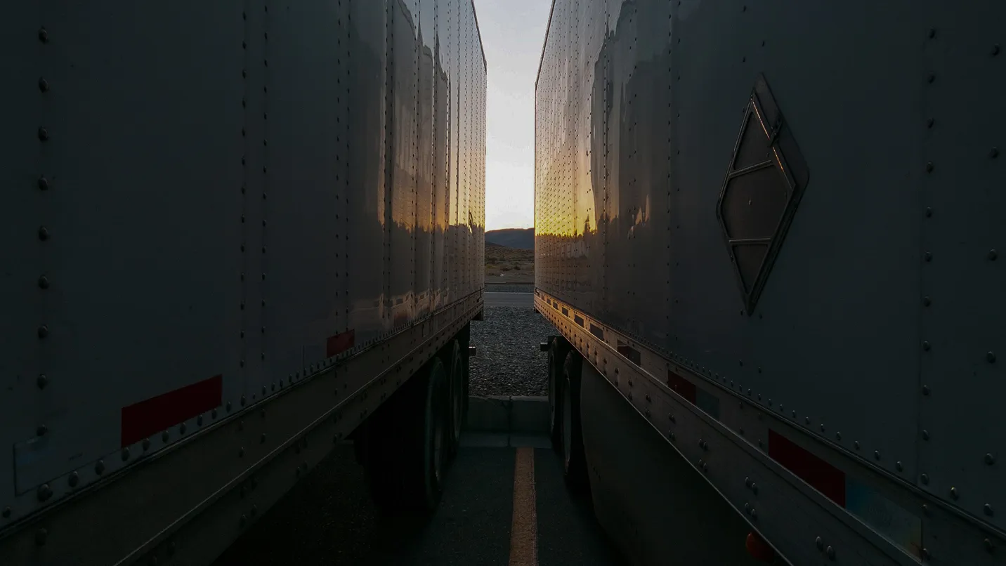 Narrow gap between two parked semi-trailer trucks at sunset with mountains in the background.