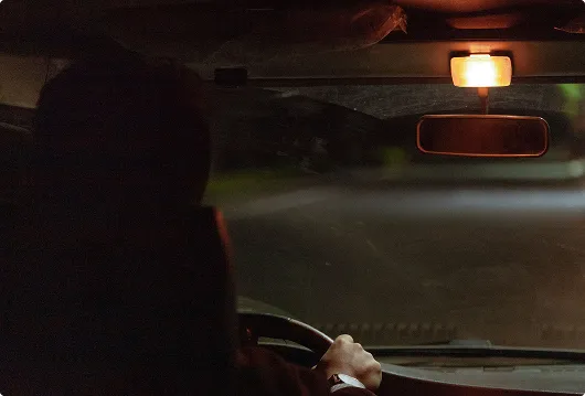 Person driving a car at night with the interior light on and rearview mirror visible.