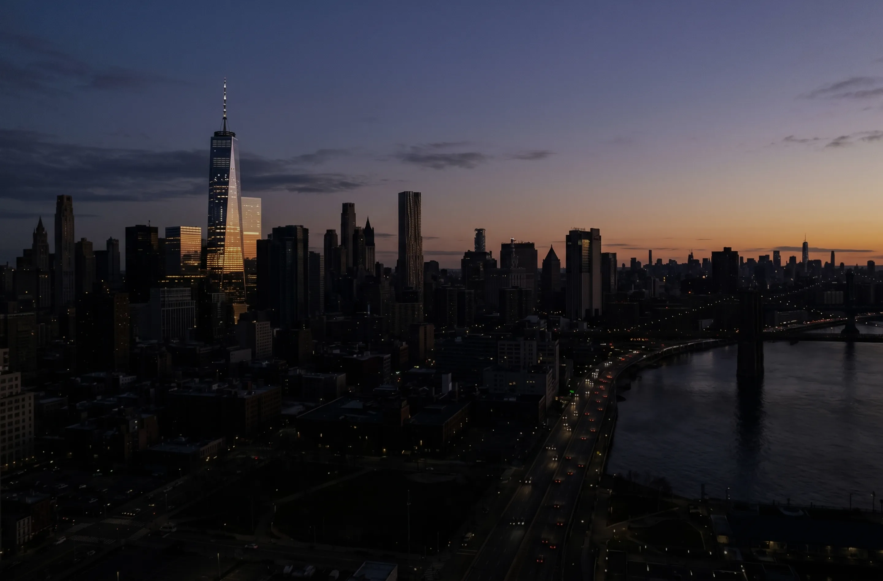 Manhattan skyline at dusk with One World Trade Center reflecting sunset light and a bridge over a river.
