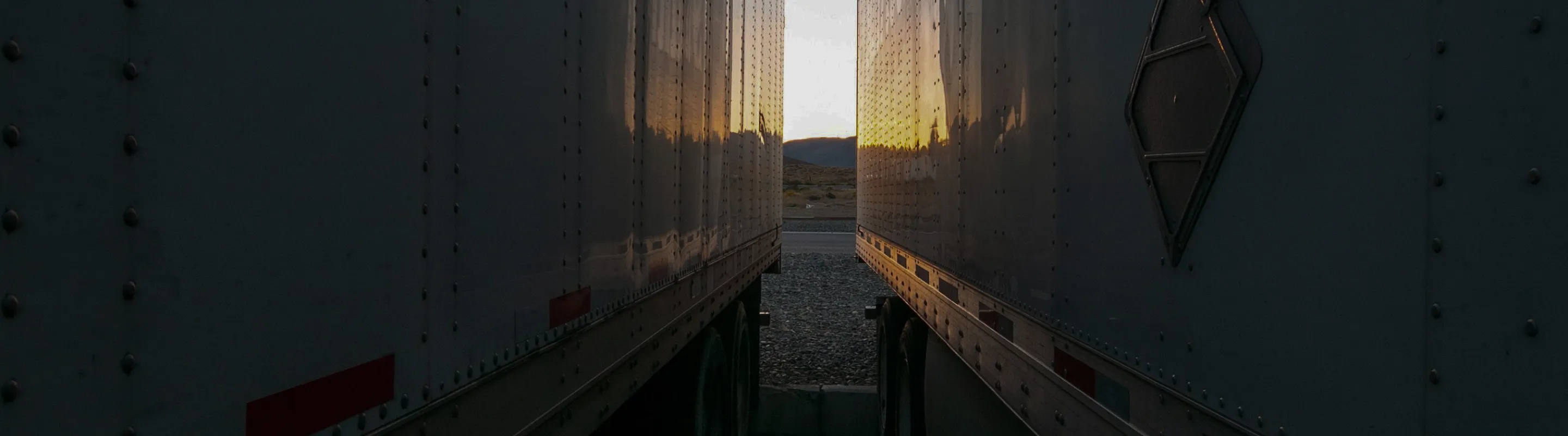 Sunset view between two parked white trailers on a gravel lot with hills in the background.