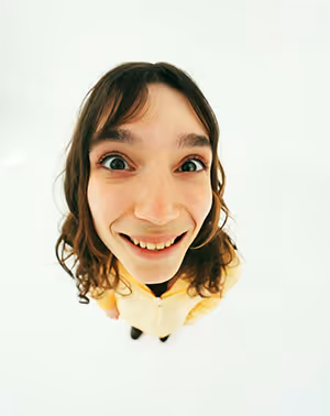 Smiling woman with curly hair looking up at the camera with a fisheye lens effect, wearing a yellow jacket.