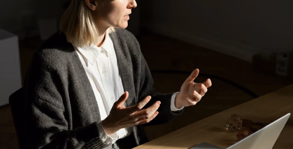 Woman in a gray cardigan and white shirt gesturing with hands while speaking, sitting at a desk with a laptop.