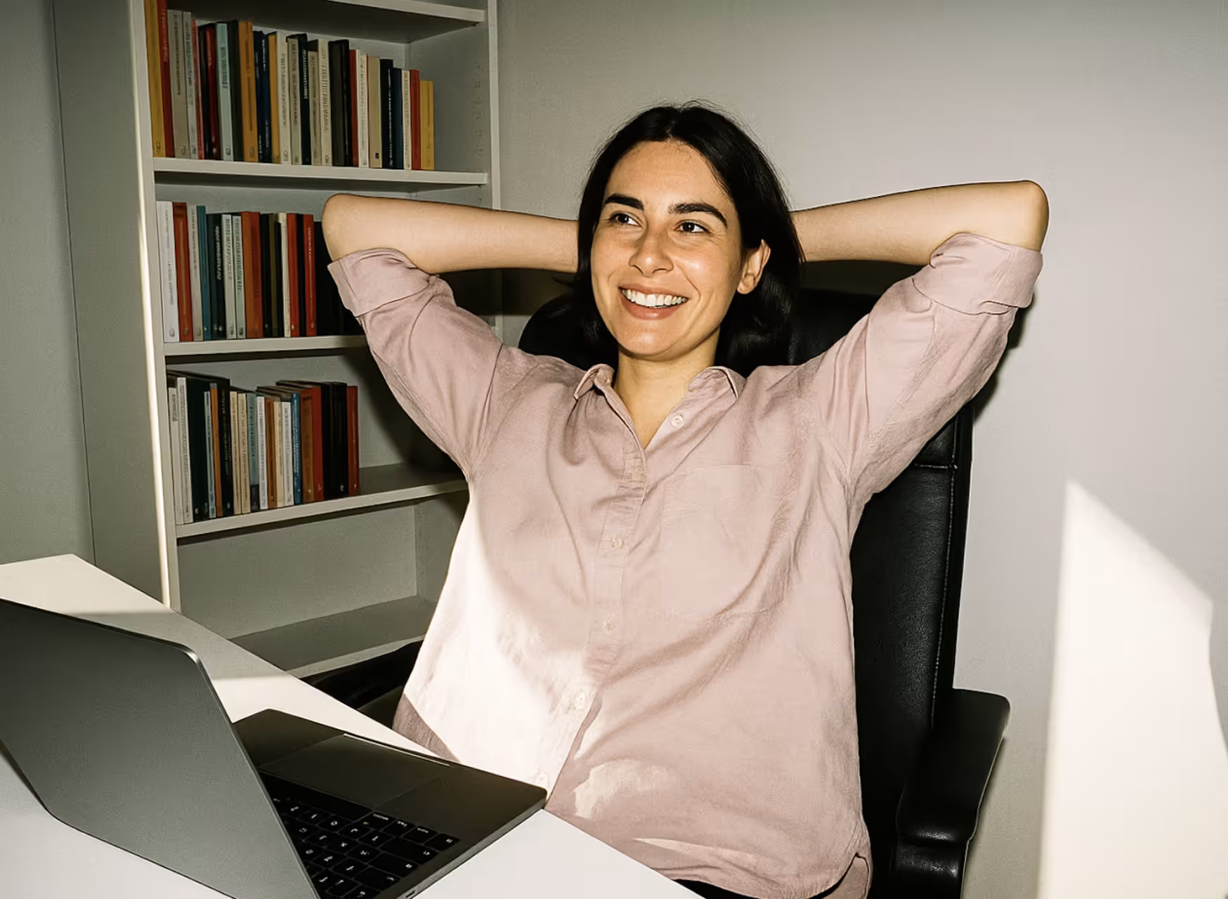 Smiling woman in a light pink shirt leaning back with hands behind her head at a desk with an open laptop.