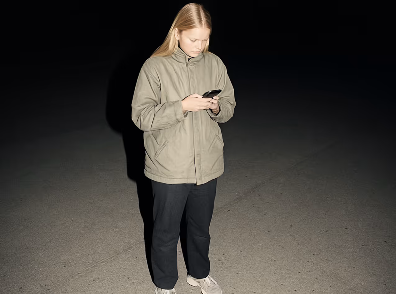 A person in a light jacket, dark pants, and white shoes standing on a pavement at night looking at a smartphone.