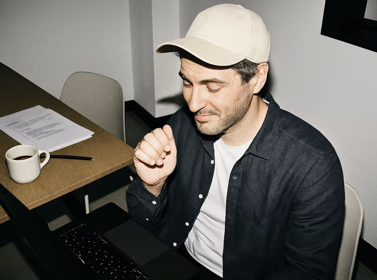 Man wearing a light beige cap and dark shirt sitting at a desk with a laptop, a cup of coffee, and papers.