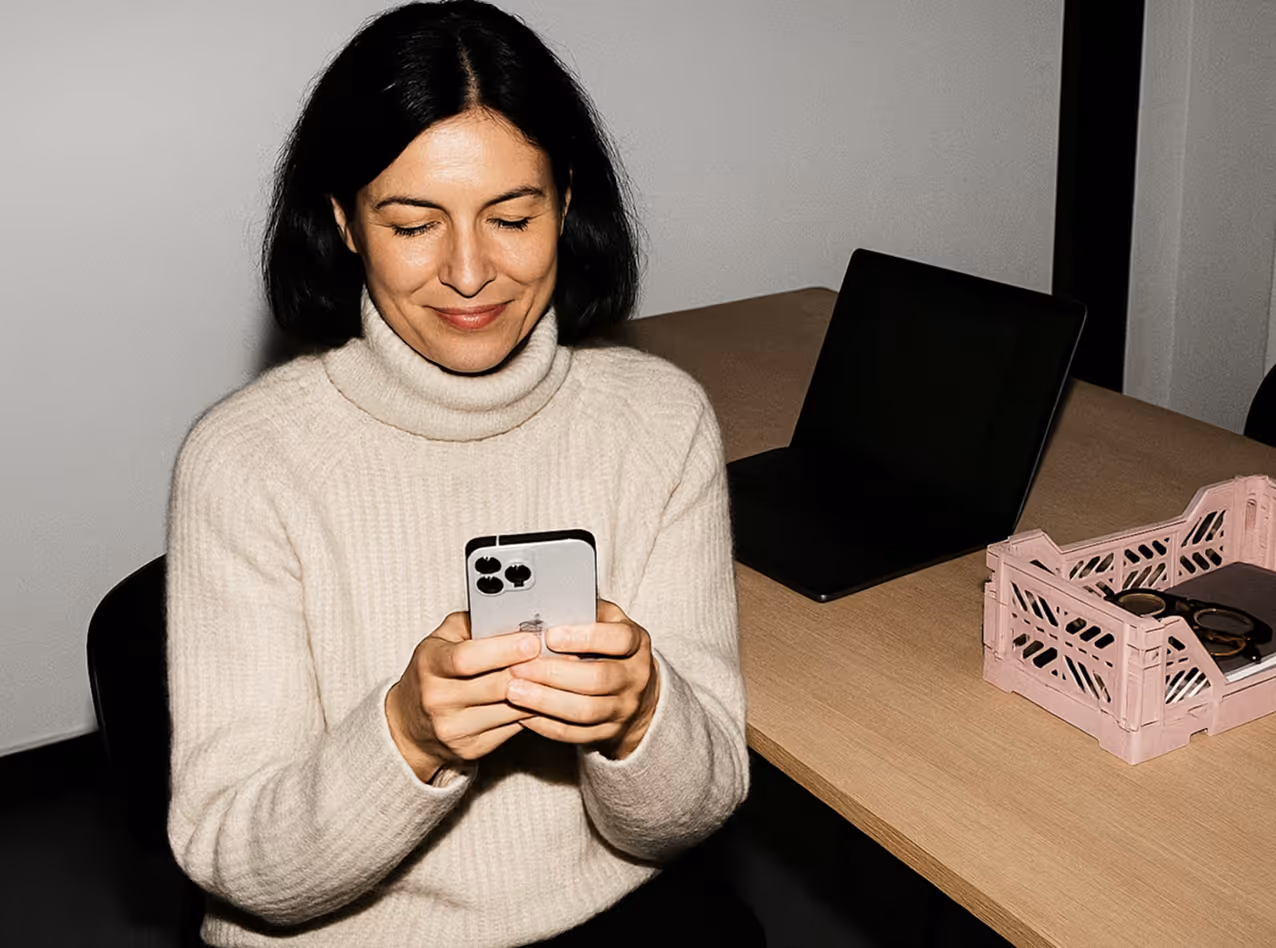 Woman in a cream turtleneck sweater smiling and looking at a smartphone while sitting at a desk with a laptop and a pink crate.