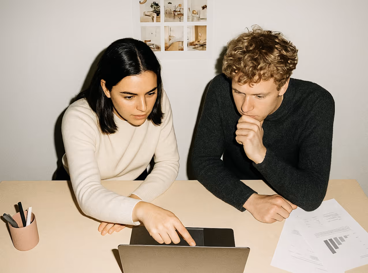 Two people sitting at a desk looking at a laptop screen, with one person pointing at the laptop and papers with charts on the desk.