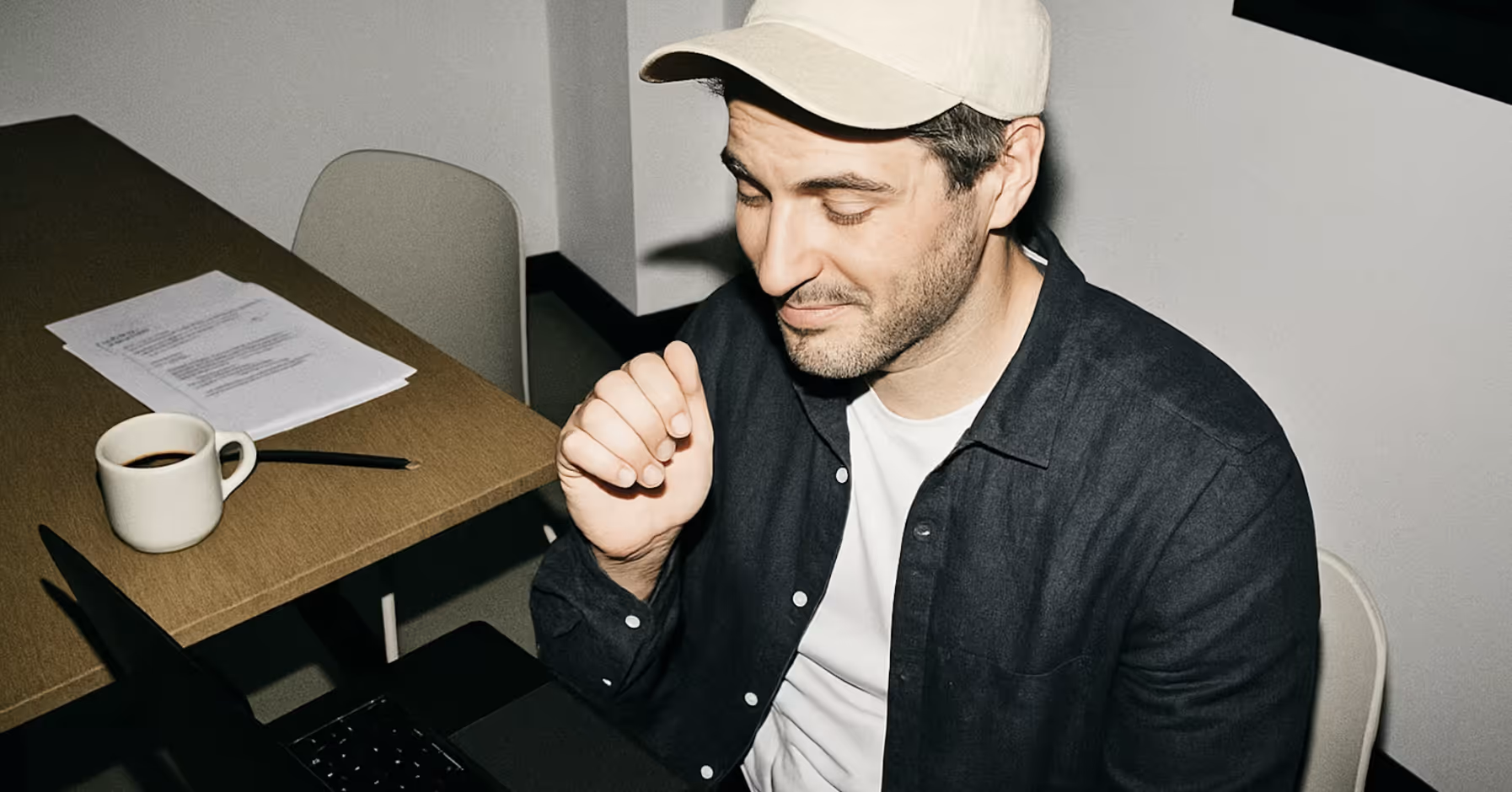 Man wearing a beige cap and dark shirt sitting at a table with a laptop, documents, a pencil, and a coffee cup.