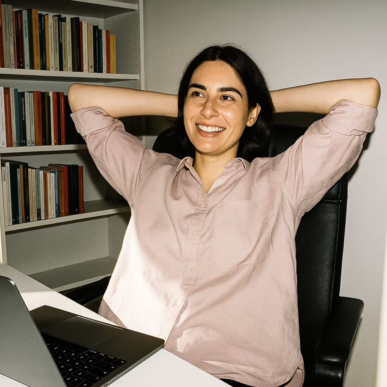 Smiling woman in a light pink shirt leaning back in a black office chair with hands behind her head near a laptop and bookshelf.