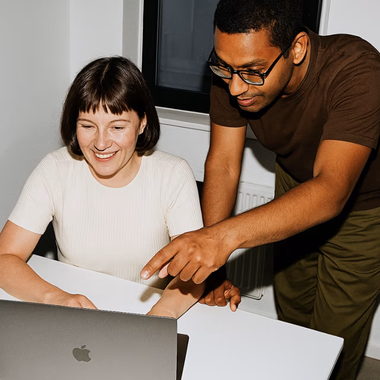 A man wearing glasses points at a laptop screen while a smiling woman looks at it.