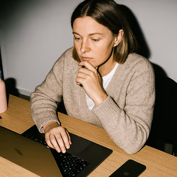 Woman in beige sweater focused on laptop screen at a wooden desk with smartphone and water bottle.