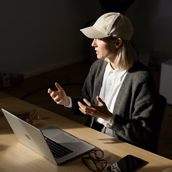 Person wearing a white cap and dark sweater speaking and gesturing with hands while sitting at a desk with a laptop, glasses, and smartphone.