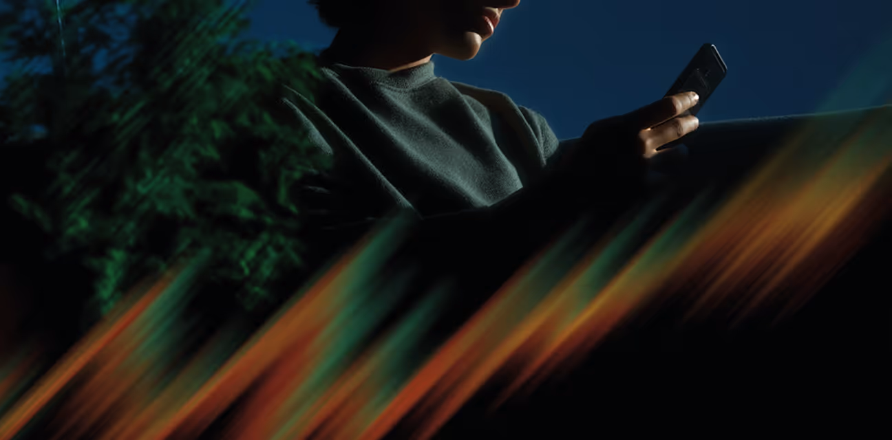 Person sitting outdoors under dim lighting, looking at a smartphone with colorful light streaks in the foreground.