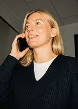 Smiling woman with blonde hair talking on a smartphone indoors, wearing a black sweater and white shirt.