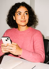 Woman with curly dark hair wearing a pink sweater, holding a white smartphone and sitting at a table with a notebook and pen.