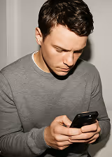 Young man in a gray sweater looking down at a smartphone he is holding with both hands.