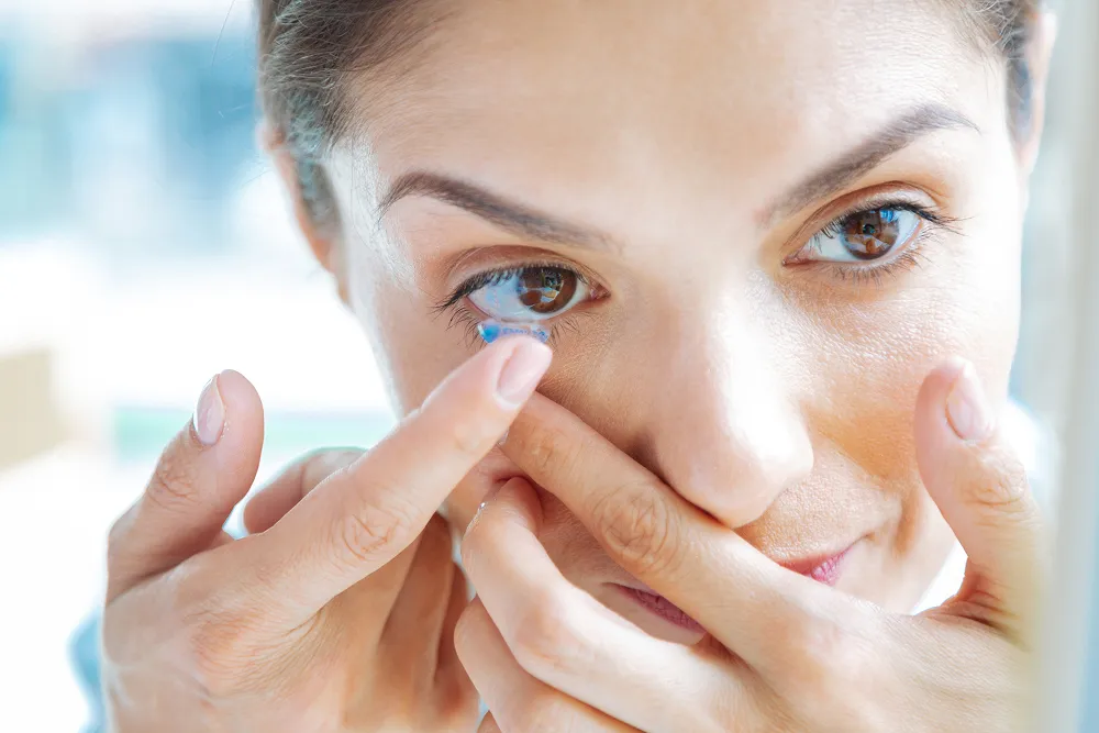 Woman inserting a blue contact lens into her right eye with both hands.
