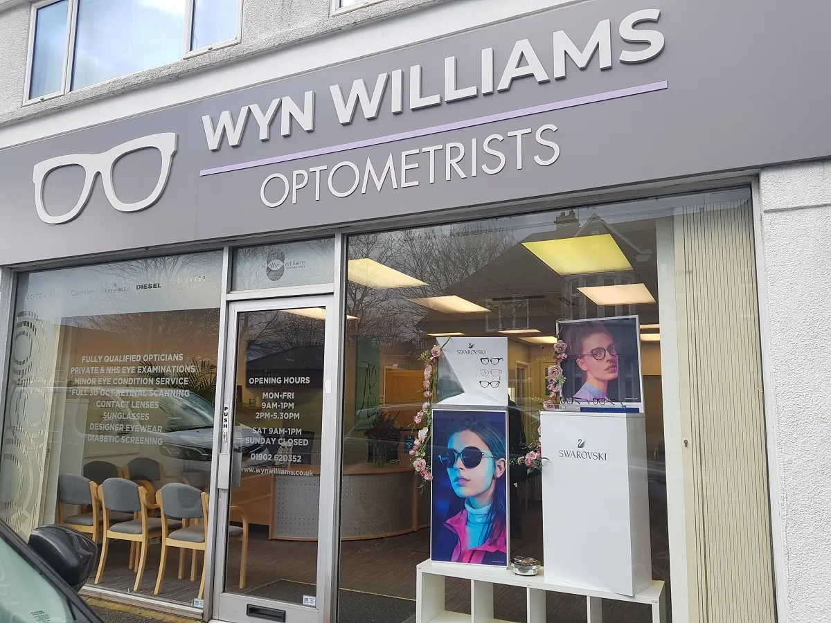 Front view of Wyn Williams Optometrists storefront with signage, glass door displaying opening hours, and window display featuring Swarovski eyewear advertisements.