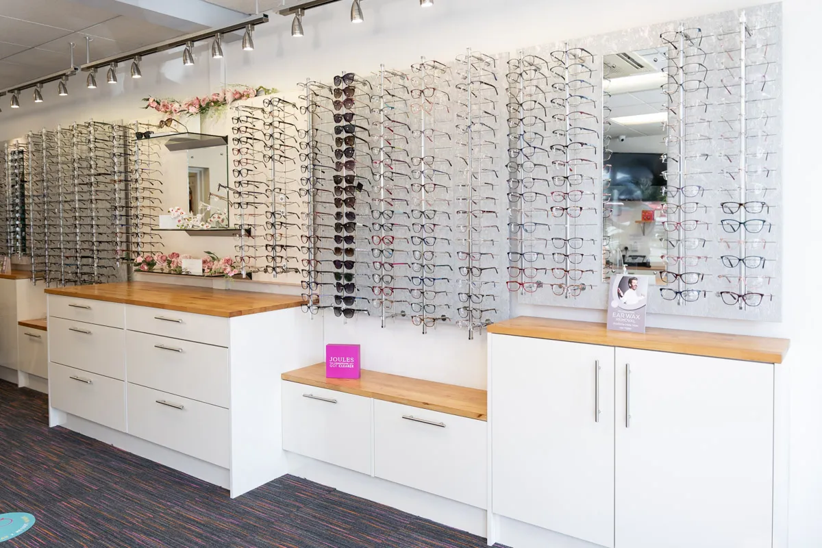 Interior of an optician store with multiple eyeglass frames displayed on wall-mounted racks above white cabinets with wooden countertops.