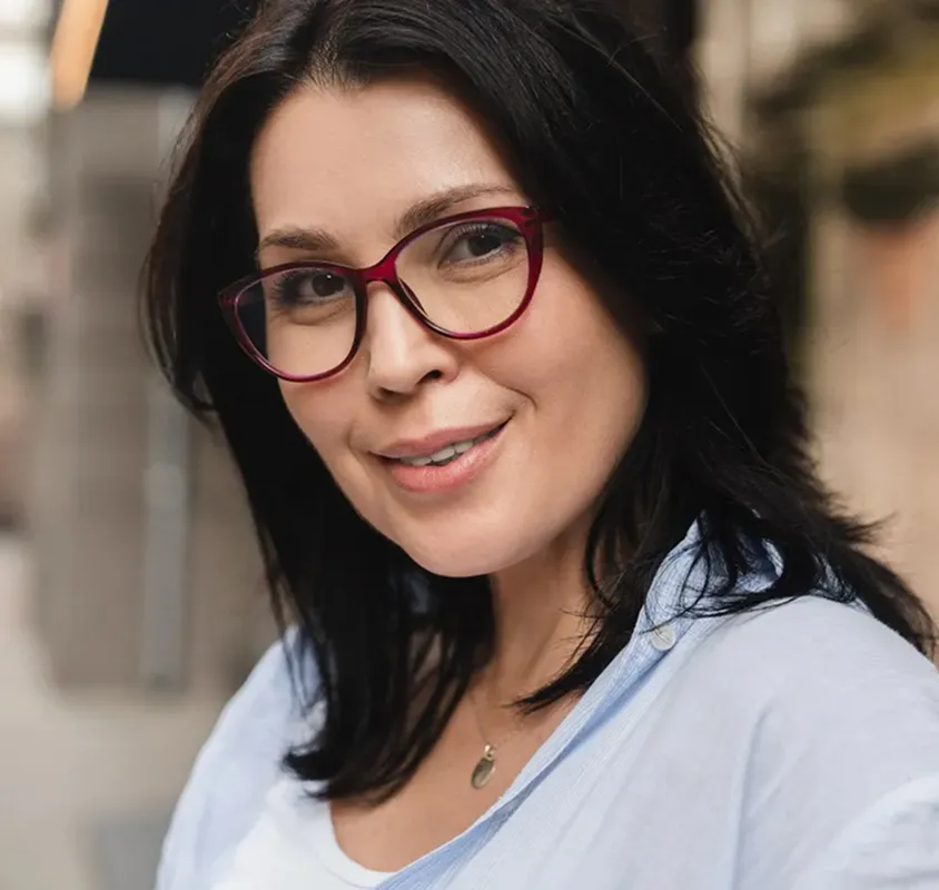 Smiling woman with dark hair and red-framed glasses wearing a light blue shirt and a necklace.