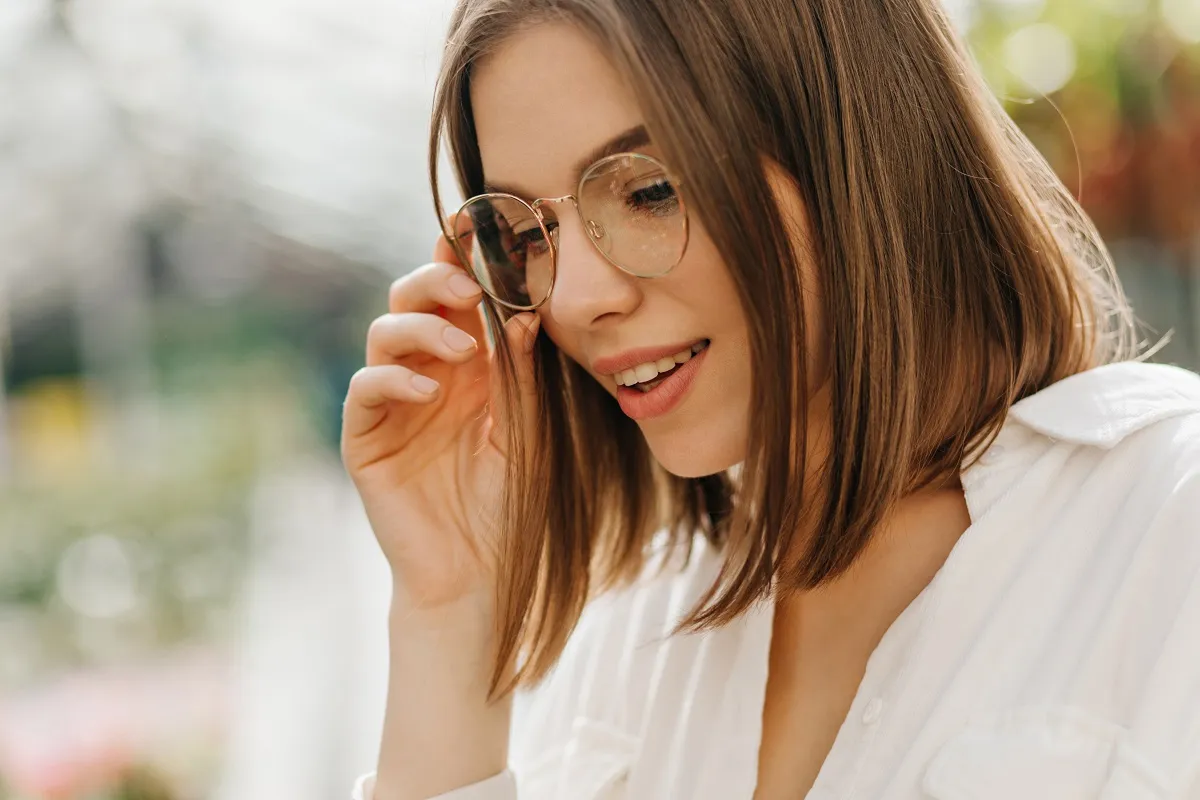 Young woman with shoulder-length brown hair adjusting her round glasses and smiling, wearing a white shirt.